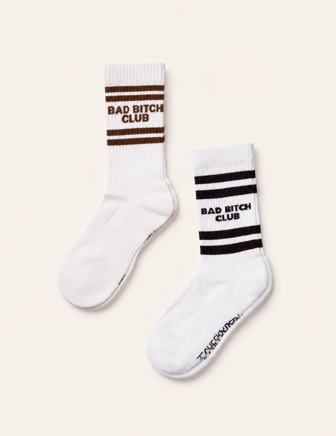 Two white crew socks with athletic stripes and the text 'BAD BITCH CLUB' on the cuff, shown on a light beige background. One sock has brown stripes and text, and the other has black stripes and text.